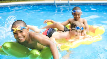 Three kids smiling and splashing in an outdoor pool while riding on pool floats